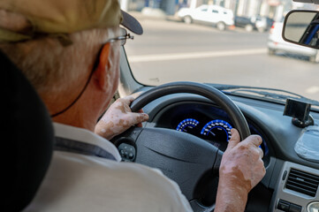 An elderly man is driving a car, holding the steering wheel with both hands, and has vitiligo on...