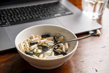 Bowl of assorted dietary supplements and capsules beside a laptop on a wooden table, illustrating health and wellness lifestyle choices
