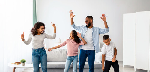 Happy Family Concept. Positive black parents dancing to music with their excited daughter and son. Playful children having fun with mom amd dad, enjoying free time with family in living room
