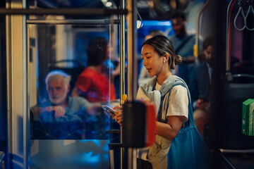 Young woman using smartphone and wearing wireless earbuds while riding public transport at night