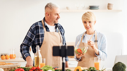 Couple cook together, explain recipe and record video for vlog and social media with smartphone camera and tripod. Happy mature wife and husband prepare food in kitchen interior, empty space, blurred