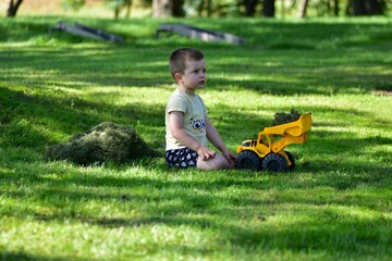 Fototapeta premium Little boy sitting on green lawn with yellow toy truck filled with grass. Summer outdoor play, childhood imagination and leisure in countryside nature, joyful and carefree moment.