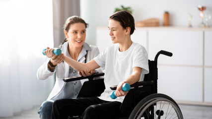 Physical rehabilitation concept. Young physiotherapist helping teenage boy in wheelchair to do...