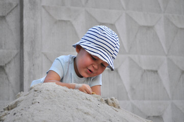 Little child in striped hat playing on a pile of sand outdoors. Toddler in light blue t-shirt and...