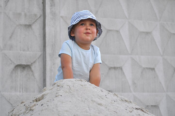 Little child in striped hat playing on a pile of sand outdoors. Toddler in light blue t-shirt and...