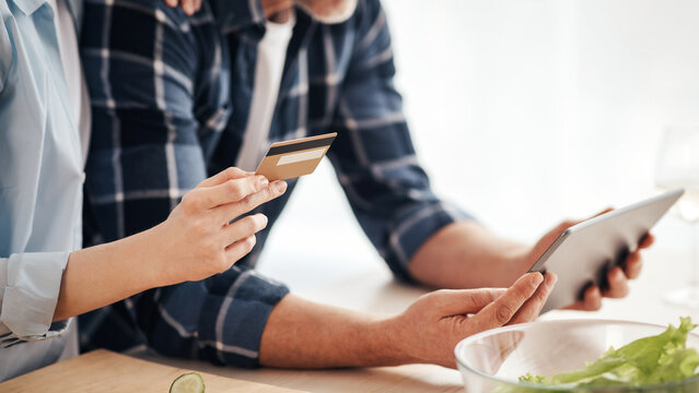 Modern ordering of groceries, fast food and shopping at home during covid-19 lockdown. Smiling senior couple look at tablet and hold credit card in kitchen interior with vegetables and salad, cropped - Powered by Adobe
