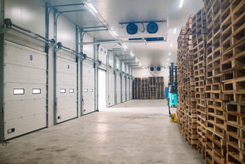 Interior of a cold storage warehouse with stacked wooden pallets, loading dock doors, and a forklift in the background. The facility is used for logistics, supply chain, frozen goods storage