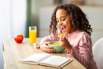 Cute black teen girl having healthy breakfast, eating cereals and drinking orange juice, preparing for lessons and reading book while sitting at desk at home