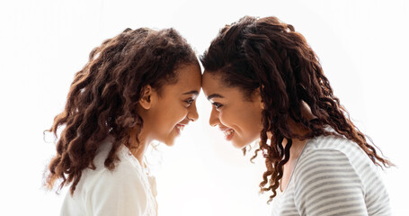 Loving african american mom and daughter touching each other with foreheads over white background, side view. Happy black young woman and school girl mother and child bonding over white