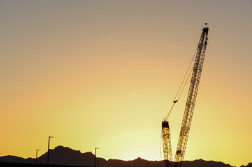 A large crawler crane stands silhouetted against a sunrise sky near a mountain range, representing heavy construction equipment used for lifting operations at infrastructure development sites