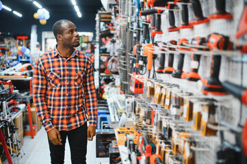 African american man holding and choosing electric screwdriver, standing in construction store