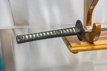 Close-up of a katana on a stand, with a ridged blade and bamboo grip, against a blurred background...