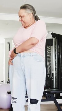 Vertical video: Senior woman entering gym tugging jeans as friend helping zip and celebrating fit