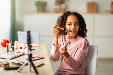 Beauty blogging. Cheerful african american teen recording cosmetics product review, showing various brushes and applying powder. Girl filming video for her channel using phone on tripod