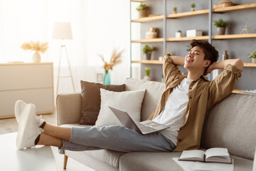 Taking Break. Smiling young asian man relaxing on comfortable couch in living room using pc laptop, happy millennial male resting on cozy sofa at home, leaning back, enjoying weekend or freelance job