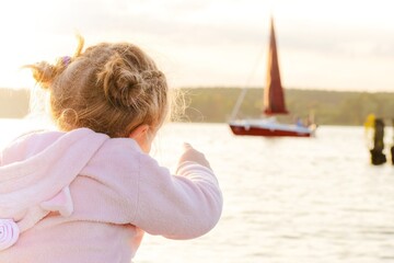 Fototapeta premium Young girl in pink hoodie points towards a sailboat on a shimmering lake during sunset, capturing a moment of wonder and exploration in a serene outdoor setting