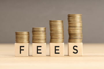 Fees word in wooden blocks with coins stacked in increasing stacks. Fees increasing concept. Copy space