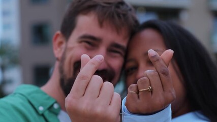 Happy multi-ethnic couple creating a korean finger heart symbol with their hands, showcasing love and affection against a vibrant urban backdrop, radiating joy and connection