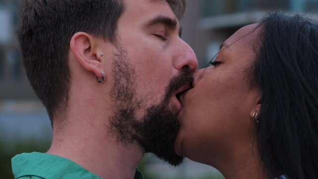 A loving interracial couple shares a passionate and intense kiss outdoors, framed by modern city buildings and greenery, capturing intimacy, affection, and urban romance.