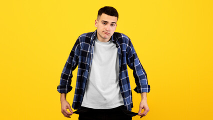 Upset young guy showing empty pockets on orange studio background. Unemployed millennial man having...