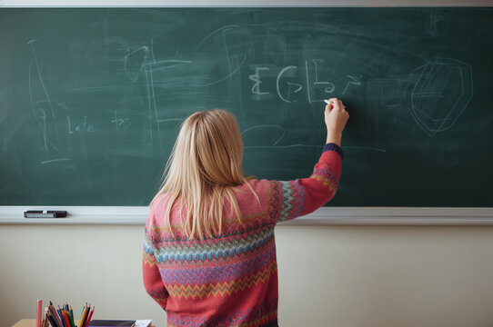 A young student with blonde hair writes on a chalkboard in a classroom setting during a lesson.
