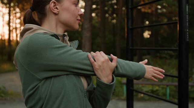 Close-up shot of young woman in sportswear stretching her shoulders before training outside in forest