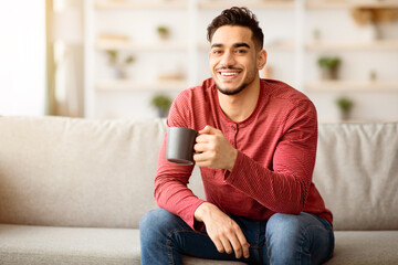 Cheerful arab guy sitting on couch and drinking tea or coffee, home interior. Happy middle-eastern...