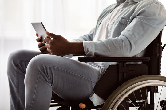 Online work and free time at home during covid-19 lockdown. Mature african american disabled guy in wheelchair, uses tablet for video call in living room or clinic interior, close up, cropped
