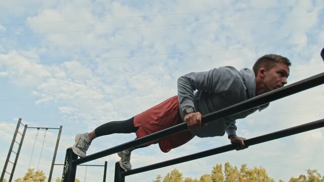 Low angle shot of young man in sportswear performing push-ups on parallel bars in outdoor training area