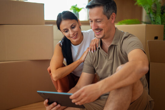 Happy couple sitting on the floor surrounded by moving boxes, looking at tablet together while planning new home, concept of relocation, family life and love.