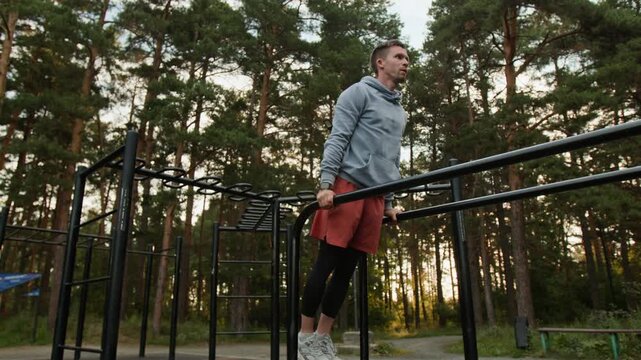 Low angle shot of young man in sportswear doing tricep dips on parallel bars in outdoor gym in forest