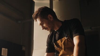 Concentrated Caucasian male with short brown hair wearing black shirt and brown apron leaning forward near coffee roasting machine. Inspecting beans. Ensuring precise roasting level for rich aroma.