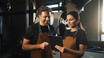 Man in brown apron gesturing with hand while standing besides woman holding tablet. Reviewing roasting data together and exchanging ideas in bright coffee production workspace. Quality control.