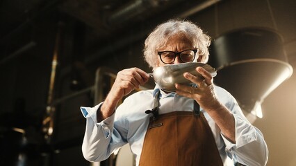 Caucasian man wearing glasses while holding metal scoop with roasted coffee beans. Smiling and looking directly. Industrial coffee roasting machine visible in bright workspace background.