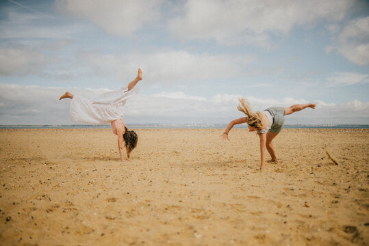 Two women are doing yoga on the beach. One of them is doing a handstand. The other woman is doing a cartwheel. The beach is sandy and the sky is cloudy