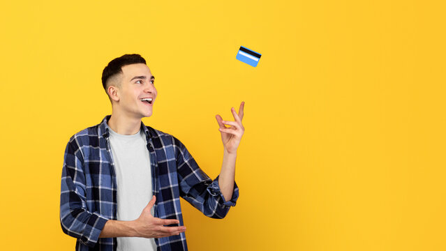 Attractive young guy throwing credit card up over orange studio background. Positive man catching bank card, using electronic money, paying contactless. Financial services concept