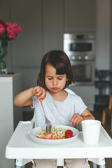 Little child girl toddler 2-3 years sitting at a table and eating pasta with salmon and cherry tomatoes in a modern kitchen, concept of childhood, vertical portrait.