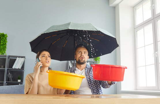 Young stressed couple sits at table under open umbrella, collecting water drips in buckets, experiencing household issue, roof leaks or plumbing problems, calling to home repair services