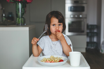 Little girl eating cherry tomato with a fork in hand sitting at the table with pasta and salmon, concept of childhood.