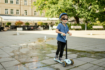 Fototapeta premium Young boy rides a scooter through a sunlit square in Vilnius, wearing a helmet and sunglasses, enjoying a warm day