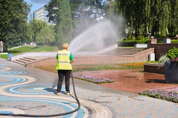 A city worker in a green vest waters flower beds on the street with a hose and a sprinkler on a...