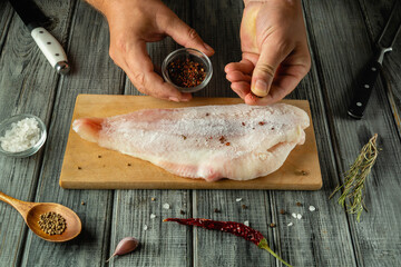 A person is adding spices to a fresh fish fillet on a wooden cutting board. Kitchen tools are...