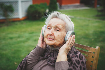 Elderly woman sitting on wooden chair outdoors listening to music through large silver headphones,...