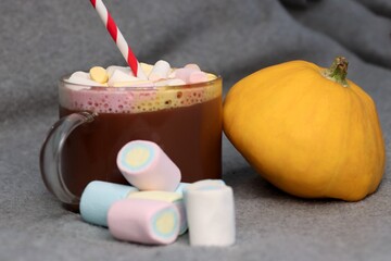 Glass of hot cocoa with marshmallows and striped straw next to bright pumpkin — cozy photo for autumn advertising campaigns, coffee shop menus, social media, seasonal greeting cards