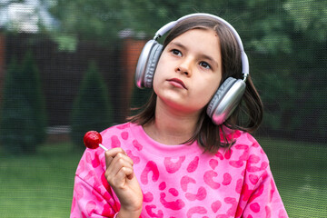 Girl in pink sweatshirt holding red lollipop and wearing headphones, listening to music with...