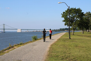 Two Joggers Running Along a Waterfront Path With a Scenic Bridge in the Background and Lush Greenery Under a Clear Blue Sky