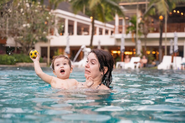 Caucasian woman holds son while swimming in pool on vacation. 