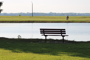 Peaceful Bench by the Lake With a Scenic View, Inviting Relaxation and Contemplation in the Afternoon Sun, Surrounded by Lush Greenery and Distant Walking Figures