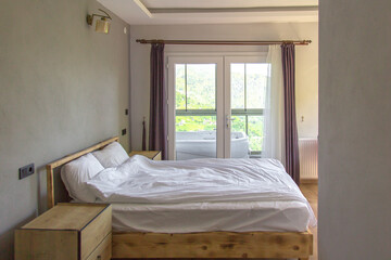 Cozy hotel bedroom with white linens, wooden furniture and a scenic mountain view through glass doors leading to a jacuzzi on the balcony, concept of travel and relaxation.