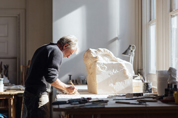 An artist meticulously carves a stone sculpture in a sunlit studio, showcasing craftsmanship, creativity, and dedication. Ideal for art, design, and creative industry concepts.